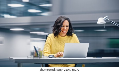 Modern Office: Happy Smiling Hispanic Businesswoman Sitting At Her Desk Working On A Laptop Computer Celebrates Victory. Latin Female Entrepreneur Is A Happy Winner. Motion Blur Background