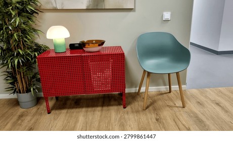 Modern office interior featuring a light-colored wall with a textured pattern. A vibrant red, perforated metal cabinet is topped with a small, frosted glass lamp, a clock, and two decorative bowls. Beside it, a sleek green chair with wooden legs stands on a warm-toned wooden floor. A potted bamboo plant adds a touch of greenery to the space.