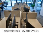 A modern office interior featuring multiple empty workstations arranged in parallel rows. Each desk is equipped with computer monitors mounted on stands and separated by beige fabric privacy dividers with visible identification numbers such as "3-B-029" and "3-B-030". The workspace features a clean, minimalist design with light-colored desktops and gray carpeting. Large floor-to-ceiling windows in the background provide natural light and reveal an urban exterior view. A single person wearing a yellow shirt and jeans is partially visible on the left side of the frame with raised arms. Office chairs, including black ergonomic models and a brown leather chair, are positioned at various stations. A potted green plant sits near white curtains on the right side, adding a touch of nature to the corporate environment. The overall atmosphere suggests a contemporary, open-plan office space designed for hot-desking or flexible working arrangements.