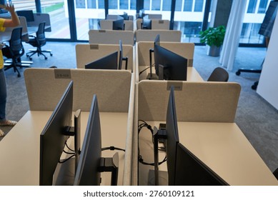 A modern office interior featuring multiple empty workstations arranged in parallel rows. Each desk is equipped with computer monitors mounted on stands and separated by beige fabric privacy dividers with visible identification numbers such as 