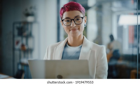 Modern Office: Portrait of Beautiful Authentic Specialist with Short Pink Hair Standing, Holding Laptop Computer, Looking at Camera, Smiling Charmingly. Working on Design, Data Analysis, Plan Strategy
