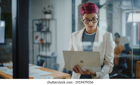 Modern Office: Portrait of Beautiful Authentic Specialist with Short Pink Hair Standing, Holding Laptop Computer, Concentrated on Her Work. Working on Design, Data Analysis, Plan Strategy - Powered by Shutterstock