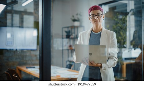 Modern Office: Portrait Of Beautiful Authentic Specialist With Short Pink Hair Standing, Holding Laptop Computer, Looking At Camera, Smiling Charmingly. Working On Design, Data Analysis, Plan Strategy