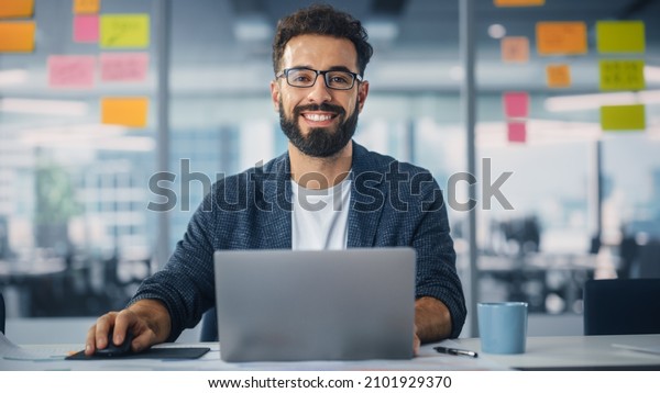 Modern Office: Portrait of Stylish Hispanic\
Businessman Works on Laptop, Does Data Analysis and Creative\
Designer, Looks at Camera and Smiles. Digital Entrepreneur Works on\
e-Commerce Startup\
Project