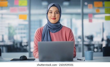 Modern Office: Portrait Of Young Muslim Businesswoman Wearing Hijab Works On Laptop, Does Data Analysis, Looks At Camera And Smiles. Empowered Digital Entrepreneur Works On E-Commerce Startup Project