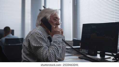 In modern office: Senior financial specialist drinks coffee, talks to business client using landline phone. PC screen shows equity market and shares. Working in International stock exchange company. - Powered by Shutterstock