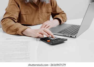En un espacio de oficina moderno, una persona con una camisa marrón se enfoca en los cálculos con una calculadora mientras escribe en una computadora portátil. Los papeles se distribuyen en la mesa cercana.