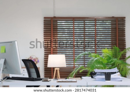 Modern office workplace setting with computer monitor and black desk chair. Showing a tidy desk with paperwork stack. Coffee cup. And a bright desk lamp