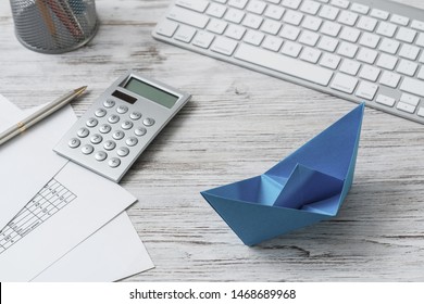 Modern Office Workspace With Blue Paper Ship. Corporate Strategy And Smart Solutions. Flat Lay Wooden Desk With Computer Keyboard, Calculator And Documents. Creative Marketing And Advertising Agency