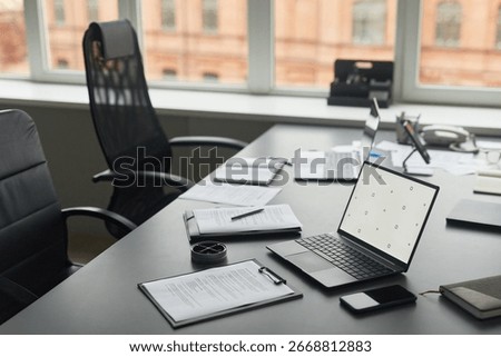 Modern office workspace featuring open laptop, smartphone, documents, pen and coffee cup on desk near large window, empty ergonomic chairs and additional laptops in background