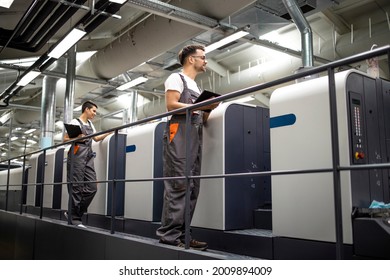Modern offset printing machine and operators in working uniform checking quality and controlling process of print.