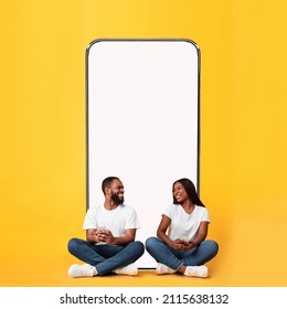 Modern Online Communication. Happy Cheerful Black Couple Sitting On Floor Near Big Giant Smartphone With White Empty Vertical Screen Using Cell Phones, Chatting On Social Media, Yellow Orange Wall - Shutterstock ID 2115638132