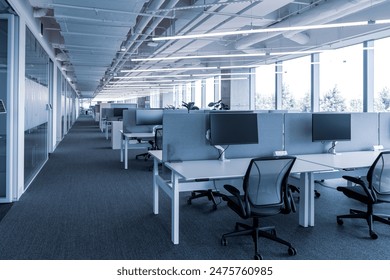 The modern open-plan office with a clean, organized layout. The workspace features rows of individual desks equipped with computers and ergonomic office chairs, separated by privacy screens. - Powered by Shutterstock