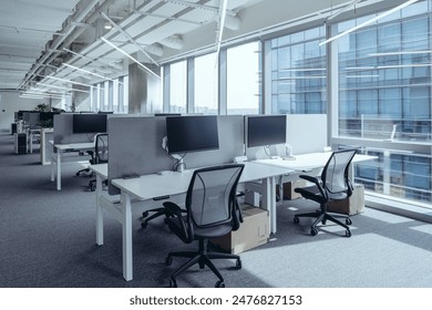 The modern open-plan office with a clean, organized layout. The workspace features rows of individual desks equipped with computers and ergonomic office chairs, separated by privacy screens. - Powered by Shutterstock