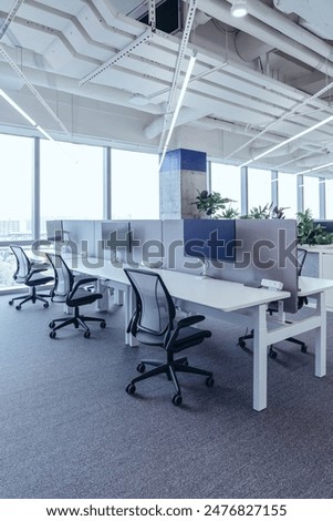 The modern open-plan office with a clean, organized layout. The workspace features rows of individual desks equipped with computers and ergonomic office chairs, separated by privacy screens.