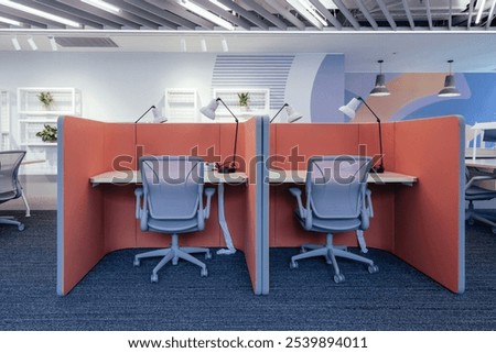 A modern open-plan office space designed for productivity and collaboration. The workspace features individual cubicles with coral-colored partitions, providing both privacy and an open environment.