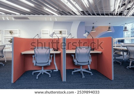 A modern open-plan office space designed for productivity and collaboration. The workspace features individual cubicles with coral-colored partitions, providing both privacy and an open environment.