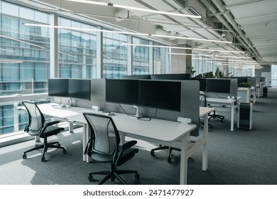 a modern, open-plan office space filled with natural light from large windows that offer a view of the cityscape. The office features a clean, minimalistic design with  chairs and computer monitors.