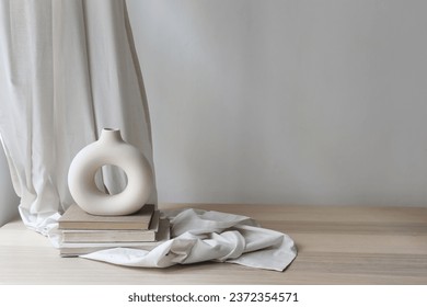 Modern organic shaped vase on old books, beige table. Linen curtain, folded drapery and empty white wall background. Aesthetic sunlight shadows. Minimalist template for home room interior product.