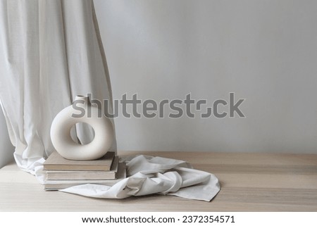 Modern organic shaped vase on old books, beige table. Linen curtain, folded drapery and empty white wall background. Aesthetic sunlight shadows. Minimalist template for home room interior product.
