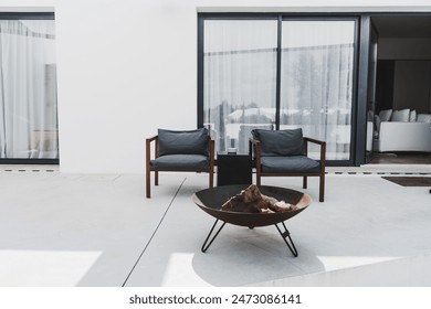 A modern outdoor patio setup: two gray cushioned wooden chairs and a round fire pit with logs. Set against a minimalistic white building with large glass sliding doors and sheer curtains - Powered by Shutterstock