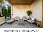 Modern outdoor patio with wooden ceiling, cushioned chairs, striped pillows, and a coffee table, surrounded by greenery.