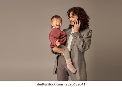 modern parenting, building successful career, empowered woman, cheerful mother talking on smartphone and holding in arms toddler daughter on grey background, motherhood, businesswoman - Powered by Shutterstock