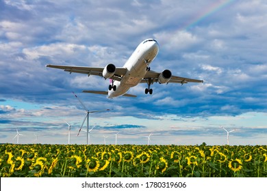 Modern passenger plane flies low over a sunflower field and wind electric generators. The aircraft is climbing against the background of a colorful sky with rainbow.