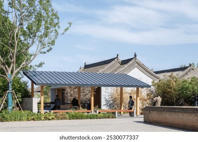 Modern pavilion with sloped roof, blending traditional and contemporary styles. Framed by trees, people interact nearby amid serene surroundings. - Powered by Shutterstock