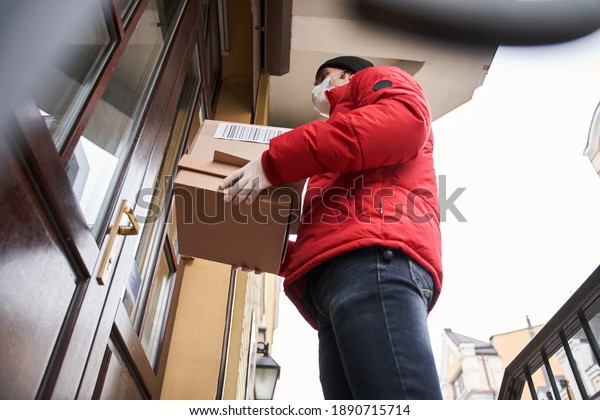 Modern post\
and delivery by car online orders. Low angle view of the courier\
wearing protective mask holding box near customer house, while\
standing near the doors. Stock\
photo