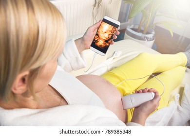 Modern Pregnant Woman Sitting On A White Couch At Home Relaxing And Doing By Herself Digital 3D Ultrasound Scan Of A Baby With A Mobile Smart Phone