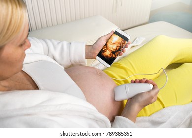 Modern Pregnant Woman Sitting On A White Couch At Home Relaxing And Doing By Herself Digital 3D Ultrasound Scan Of A Baby With A Mobile Smart Phone