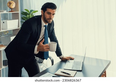 Modern professional businessman at modern office desk using laptop to work with coffee in his hand. Diligent office worker working on computer notebook in his office work space. fervent - Shutterstock ID 2306826367