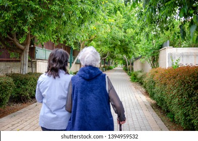 Modern Psychotherapist Prefers Working With An Elderly Lady In An Informal Setting. They Walking Away In A Green Alley From The Camera. Their Backs Are Not In Focus.