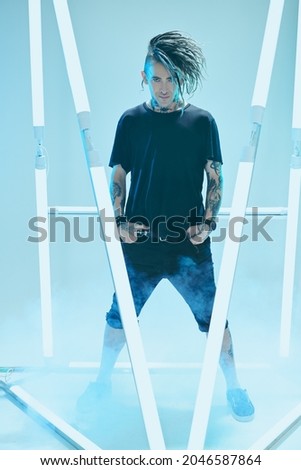 Modern punk rock musician with dreadlocks and in black clothes  posing among neon lamps. Space punk rock music. Youth alternative culture. Full length portrait.
