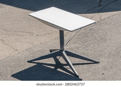 Modern rectangular metal outdoor table featuring star-shaped legs, clean minimalist design, light-colored laminate surface, cylindrical metallic base column, textured paved setting, daytime lighting.