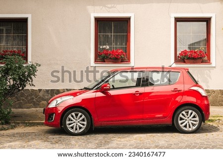 Modern red car parked on slope outside an old house with red windows and flowers, car on the old european street.
