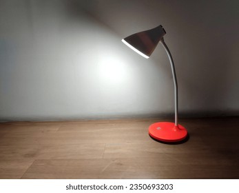 A modern red desk lamp with a white curved neck, powered by rechargeable batteries, emits soft LED light onto a wooden surface, creating a minimalist, cozy atmosphere. - Powered by Shutterstock