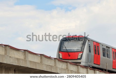 Modern red and silver metro train driving on a concrete elevated viaduct against a cloudy sky.