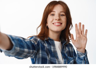 Modern redhead girl video chatting, waving hand at smartphone camera and smiling, taking selfie, record blog on phone, holding device with extended hand, say hi, standing over white background