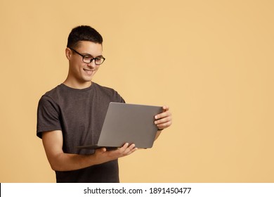 Modern Remote Communication Online, Stay At Home, During Covid-19 Lockdown. Smiling Millennial Guy Student With Glasses Looks At Laptop And Works On Project, Isolated On Beige Background, Studio Shot