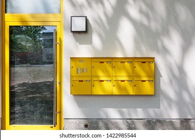 Modern Residential Apartment Building Entrance With Glass Door, Yellow Mailboxes And Tree Shadow On Wall. New Home Currently Under Construction
