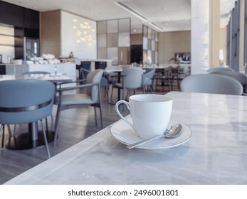 Modern café or restaurant interior, focusing on a white coffee cup with a saucer and spoon placed on a marble table. The background features elegant, contemporary seating with gray upholstered chairs  - Powered by Shutterstock