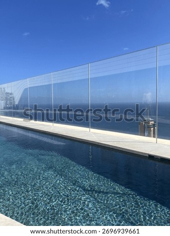 Modern rooftop infinity pool with clear blue water reflecting the sky, surrounded by glass panels overlooking a vast ocean horizon under bright sunlight and a clear blue sky, serene atmosphere.