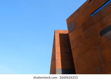 A modern rust-colored building with sharp geometric lines stands out against a clear blue sky.