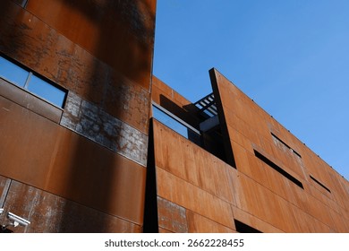 A modern rust-colored building with sharp geometric lines stands out against a clear blue sky