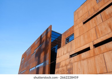 A modern rust-colored building with sharp geometric lines stands out against a clear blue sky