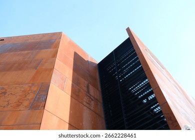 A modern rust-colored building with sharp geometric lines stands out against a clear blue sky