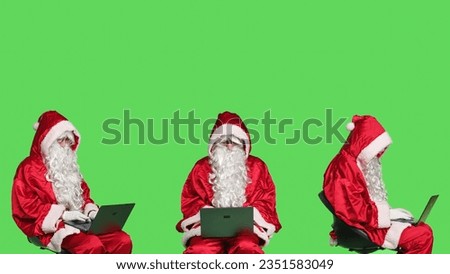 Modern santa claus works on laptop, greenscreen backdrop in studio. Father christmas sitting on chair and using wireless pc for online website browsing, searching for children addresses.