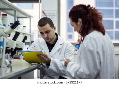 Modern scientist working with pipette in biotechnology laboratory equipment for research on a white table and an assistant who records the measured results